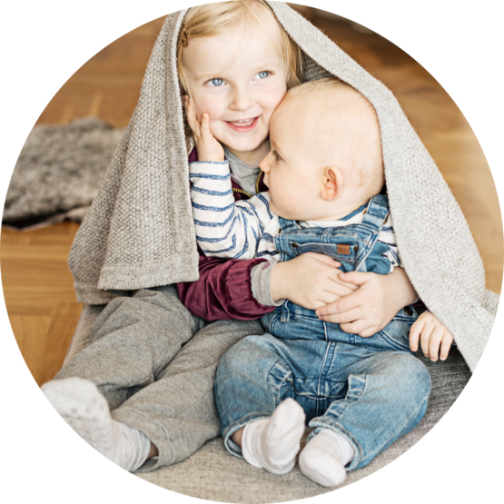 Two young siblings sitting on the floor, with an older child in a gray outfit hugging a baby in denim overalls, both covered by a cozy gray blanket.