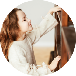 A young girl with long brown hair is reaching up to touch a shelf. She is wearing a cream-colored sweater with decorative bows on the sleeves