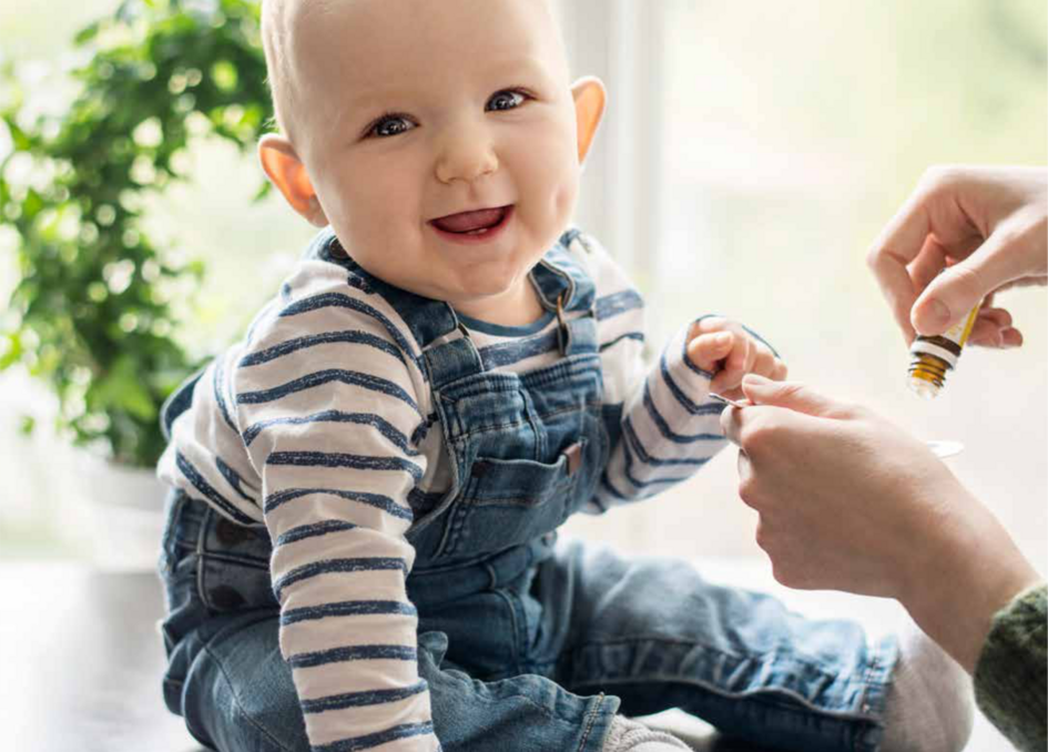 Smiling baby in a striped shirt and denim overalls sitting on a table, with an adult administering drops from a small bottle.