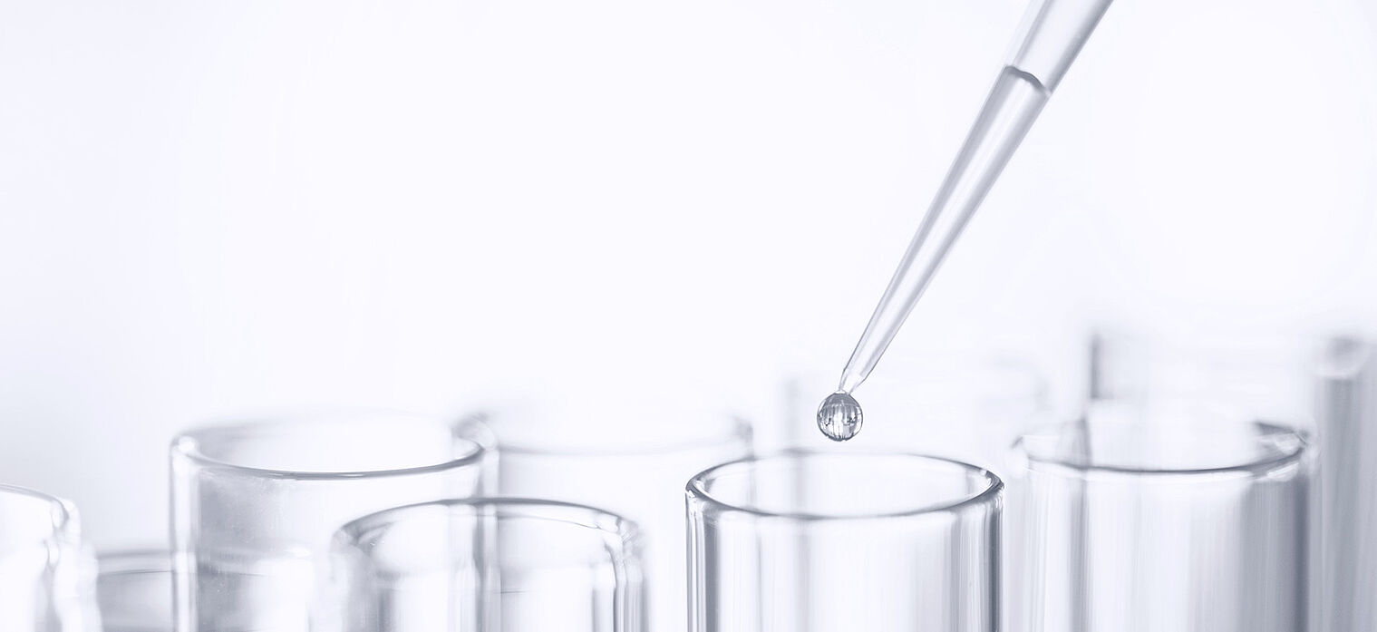 A close-up of a pipette dropping a liquid into a row of clear glass test tubes in a laboratory setting.