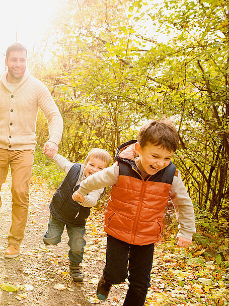 Smiling father holding hands with two young boys as they run along a sunlit forest path in autumn, with colorful leaves around them