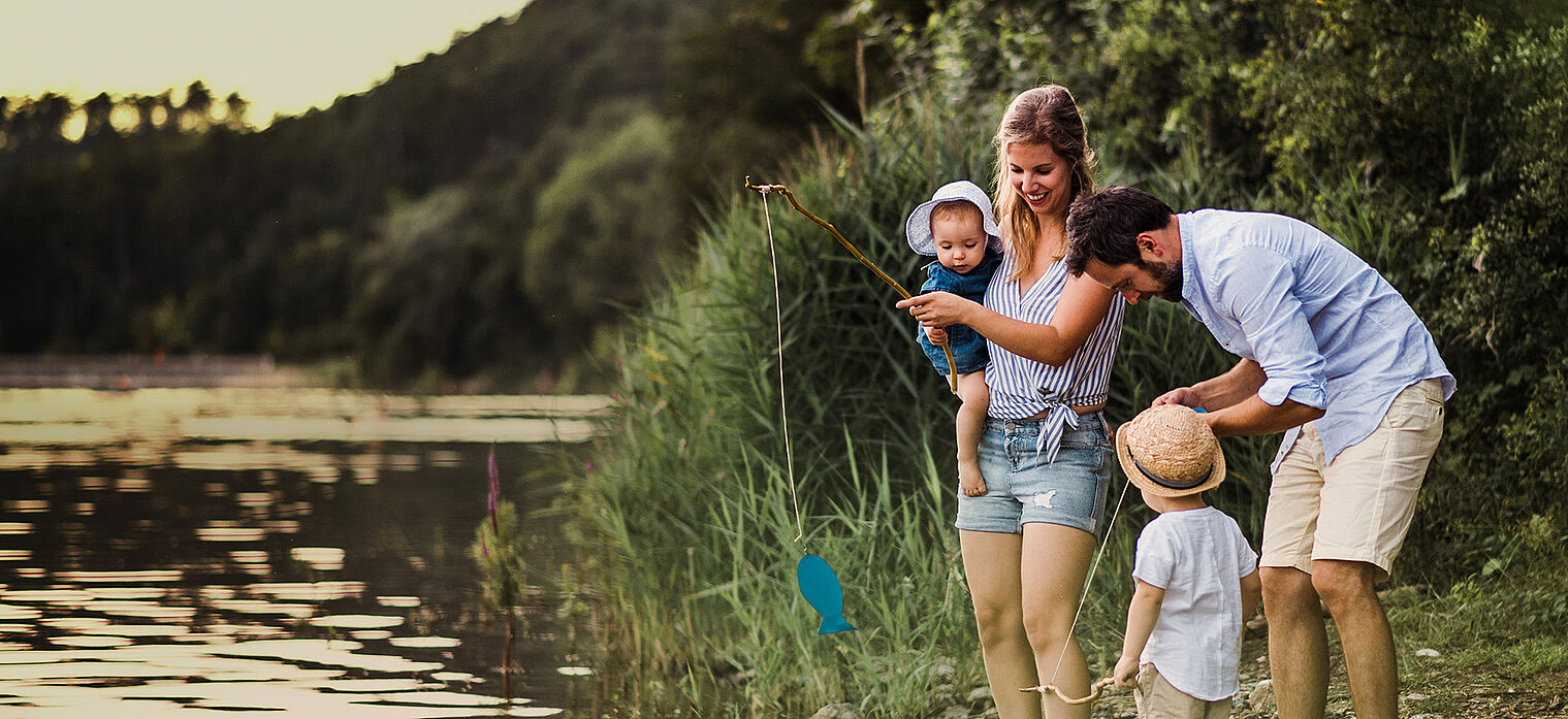 A family of four by a lake, with the mother holding a baby and helping an older child fish with a net, and the father assisting the child. They are surrounded by lush greenery.